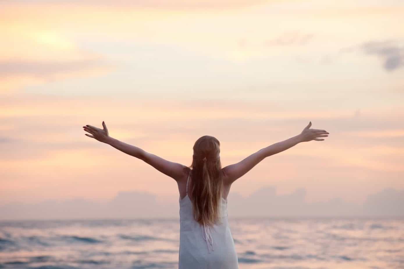 Quotes to motivate you to declutter your life | rear view of woman with arms raised at the beach during sunset